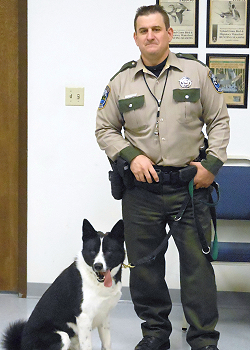 Photo of Dave Jones in uniform standing next to K9 partner Indy in WDFW department office.